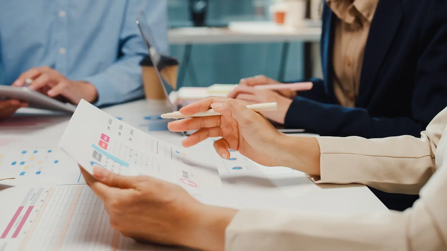 3 people sitting at a table, one typing on a laptop, one holding a tablet, and one writing on a piece of paper with graphs